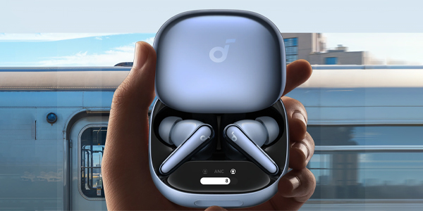 Blue wireless earbud case open in a hand, earbuds visible inside, with a blurred transit background behind it.