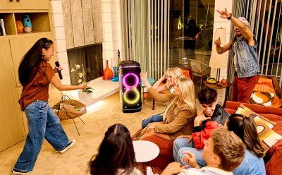 Woman singing into a microphone at a cozy living-room party while friends watch a neon-lit speaker center stage in the room.