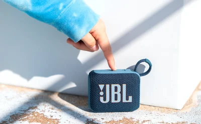 Blue JBL portable Bluetooth speaker on a textured concrete surface with a finger pressing the top button on the device.