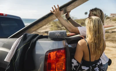 Two people load a long surfboard onto the back of a gray pickup at the beach, with a JBL speaker resting on the tailgate and clothing draped over the side of the bed.