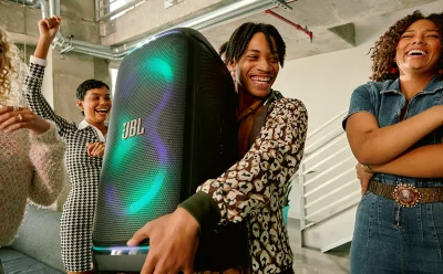 Group of diverse friends laughing and dancing around a large JBL speaker in a bright modern space.