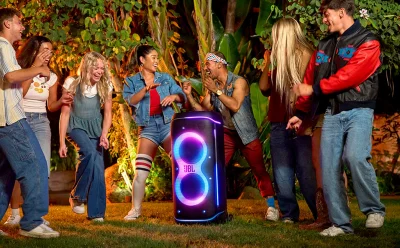 Group of friends dancing and laughing around a glowing JBL speaker in a backyard party scene at night.