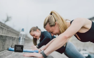Two women exercise on outdoor steps, leaning into a forward push-up; a JBL speaker, water bottle and towel sit on the bench nearby.