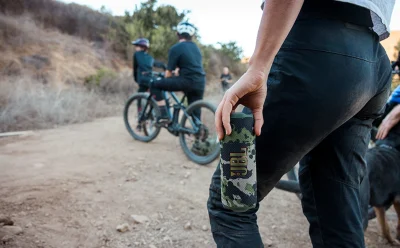 Close-up of a camo water bottle in a rider’s hand on a dirt mountain-biking trail, with cyclists and a dog in the background