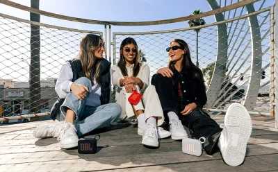 Three women sit on a rooftop deck, smiling and chatting, with small JBL speakers placed near their feet for a casual outdoor gathering.