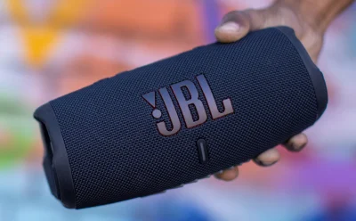 Portable JBL Bluetooth speaker being held in a hand, underscored logo visible on its fabric grille.