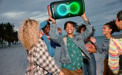 Group of diverse friends dancing on a beach, raising a large glowing speaker above their heads.