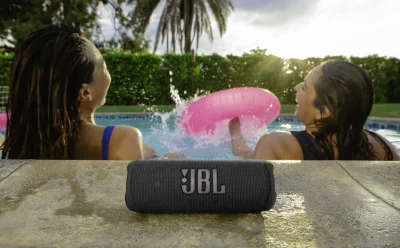 Black JBL portable speaker resting on a wet pool deck with two women swimming in the background.