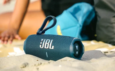Blue JBL Bluetooth speaker resting on sandy beach with a turquoise backpack blurred in the background