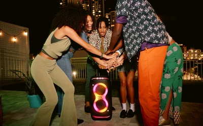 Group of friends in colorful outfits circle around a glowing JBL speaker on a rooftop at night, hands joined in a cheer.