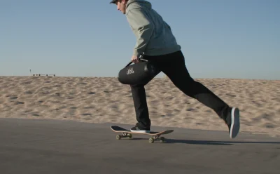Skateboarder gliding along a beach boardwalk, carrying a cylindrical JBL speaker bag under his arm.",