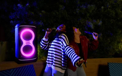 Two people singing into wireless microphones beside a glowing JBL speaker at night on an outdoor patio.