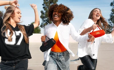 Three friends dancing outdoors holding JBL speaker bags with big smiles on a sunny day.