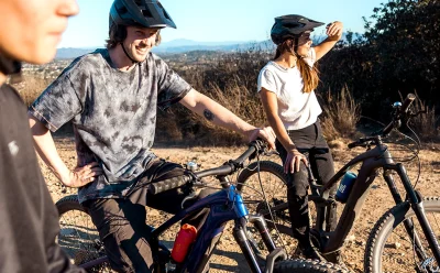 Two cyclists wearing helmets stand with their mountain bikes on a dry, rocky trail, smiling and chatting under a clear sky.