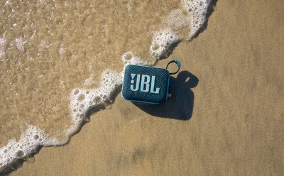 Blue JBL portable Bluetooth speaker on wet sand near the foamy shoreline.”,