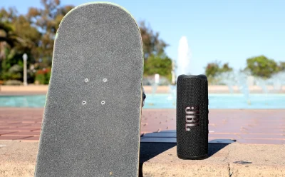 Skateboard standing upright beside a black JBL speaker outdoors near a poolside brick surface.