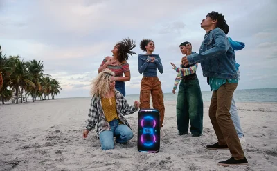 Group of diverse friends dancing on a sandy beach with a glowing JBL speaker in the foreground.