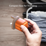 Hand holds a small orange travel plug adapter with two flat blades, suitcase in the background, illustrating compact size for traveling.