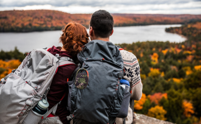 Two hikers with backpacks stand on a cliff overlooking a lake and colorful autumn forest from behind.