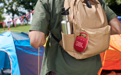 Back view of a person in a green shirt wearing a tan backpack at a campsite; a water bottle sits in a side pocket and a red JBL speaker is clipped to the bag's front pocket with tents in the background.