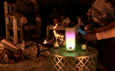 Outdoor evening scene around a fire pit with a glowing JBL speaker lantern on a metal table, while someone uses a smartphone nearby.