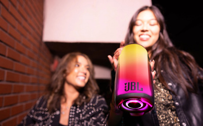 Two women in sparkly outfits posing with a rainbow JBL microphone at a party, brick wall in the background.
