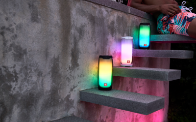 Three JBL portable speakers with rainbow LED lights placed on outdoor concrete stairs, with a person sitting at the top edge nearby.