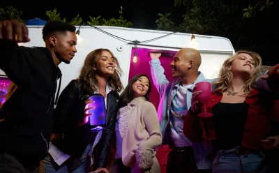 Group of friends dancing and posing with drinks near a lit trailer at night, having a good time together.