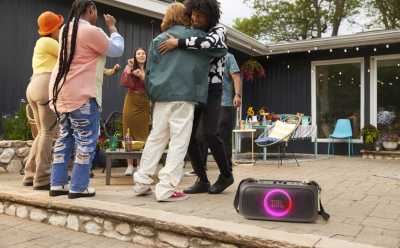 Group of friends dancing at an outdoor backyard party, with a glowing pink JBL speaker in the foreground and string lights overhead.