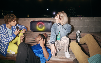 Three friends sit on a wooden bench at night, gathered around a JBL speaker with a glowing ring light between them.