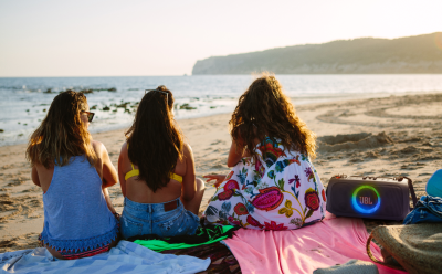 Three friends sit on a sandy beach facing the ocean at sunset, with a colorful JBL speaker nearby to the right.