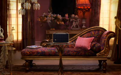 Ornate living room with a dark blue JBL speaker on a glass coffee table in front of a patterned upholstered sofa with pink pillows, illuminated by warm ambient light and vintage decor in the background.