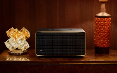 Portable JBL speaker with black front and gold trim on a wooden shelf, flanked by decorative lamps on each side.