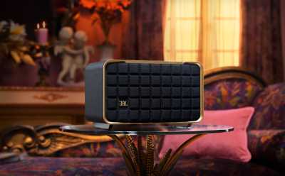 JBL portable speaker with a quilted black grille and gold trim on a glass table in a warmly lit living room.