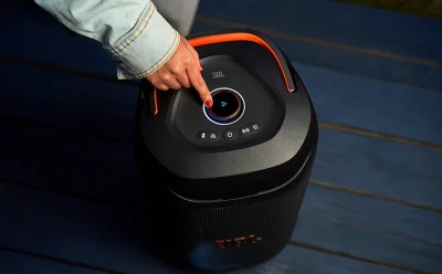 Person pressing the circular power/play button on a black JBL portable speaker with an orange handle.