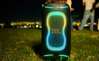 Portable JBL Bluetooth speaker on grass at night, illuminated by green and blue LED rings with a city lights backdrop behind it.