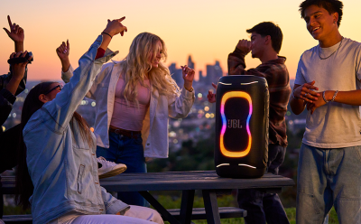 Group of friends dancing around a glowing JBL portable speaker on a picnic table at sunset with a city skyline in the background
